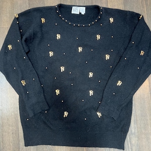 Könnte beinhalten: Schwarzer Pullover mit goldfarbenen Schleifen und Punkten. Der Rundhalsausschnitt ist mit kleinen goldenen Perlen verziert. Der langärmlige Pullover hat einen gerippten Saum und Bündchen. Der Markenname "HE RO" ist oben am Rücken sichtbar.