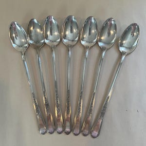 May include: Seven silver spoons arranged in a row. The spoons have a classic design with elongated bowls and slender handles. The handles feature a decorative floral pattern near the base. The spoons are reflective, showing the surrounding environment.