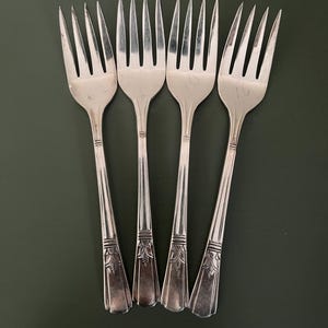 May include: Four vintage silver forks on a dark green surface. Each fork has four tines and a decorative floral pattern on the handle. The forks are made of a shiny, reflective metal.