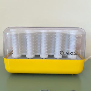 Op de afbeelding: Een vintage Clairol haarroller set. De basis is felgeel, met een doorzichtige plastic deksel. Binnenin zitten witte rollers. Het Clairol-logo is op de voorkant gedrukt. Het netsnoer is zichtbaar.