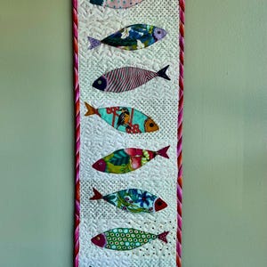 May include: A vertical quilted wall hanging featuring seven colorful fish appliqués. Each fish has a unique patterned fabric body, with red or black fins and eyes. The background is white with small black dots.