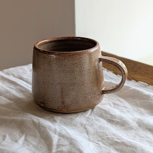 May include: A speckled brown ceramic mug with a cylindrical body and a curved handle. The exterior features a textured, earthy glaze with darker brown flecks throughout, while the rim shows a slightly darker, reddish-brown tone.