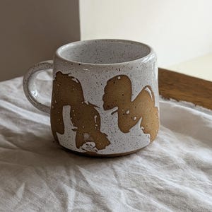 May include: A ceramic mug with a speckled white glaze and two large, abstract brown shapes painted on the exterior. The mug features a rounded body, a sturdy handle on the left side, and a smooth, curved rim. The base of the mug is brown.