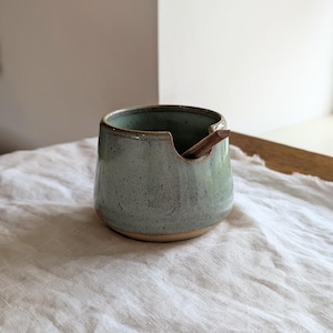 May include: A small, round, ceramic jar with a speckled light green and blue glaze. The jar features a wide opening with a small notch on the rim, designed to hold a thin, dark brown wooden spoon that rests inside the vessel at an angle.