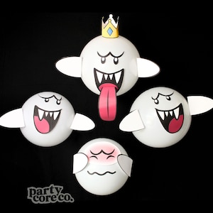 May include: Four white spherical decorations featuring cartoon ghost faces with black outlines and white teeth. One ghost wears a yellow crown and has a long pink tongue. Another ghost has pink cheeks and closed eyes. The text party core co. is visible.