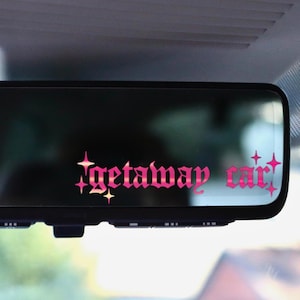 Peut inclure: Rétroviseur de voiture avec le texte "getaway car" en rose avec des étoiles. Le miroir est rectangulaire et monté à l'intérieur d'un véhicule.