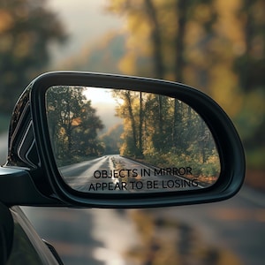 May include: A car's side view mirror with the text "Objects in mirror appear to be losing" printed on the bottom of the mirror. The mirror reflects a road with trees on either side.