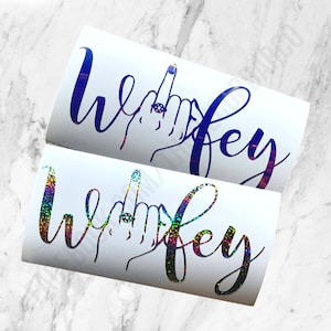 Peut inclure: Deux autocollants en vinyle avec le mot "wifey" en écriture cursive. Un autocollant est bleu et l'autre est iridescent. Les deux autocollants présentent une main avec une bague au doigt.