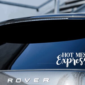 May include: A white vinyl decal with the text "HOT MESS Express" in a cursive font, applied to the rear window of a silver car.