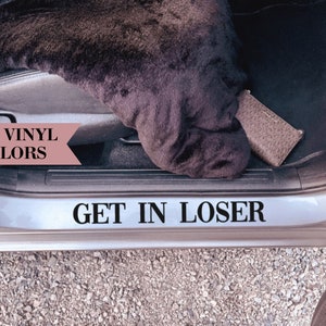 Op de afbeelding: Een zilveren autodeur met een zwarte vinyl sticker waarop "GET IN LOSER" staat. De sticker zit aan de onderkant van de deur. In de linkerbovenhoek van de afbeelding is een roze banner te zien met de tekst "30+ VINYL COLORS".