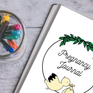 Pode incluir: Um Di&aacute;rio de Gravidez branco com uma ilustra&ccedil;&atilde;o de uma cegonha carregando um beb&ecirc;. O di&aacute;rio tem uma borda circular preta com folhas verdes no topo. Um frasco de vidro com marcadores coloridos &eacute; vis&iacute;vel no fundo.