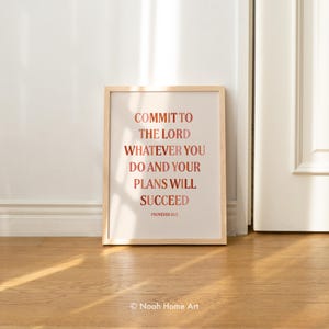 May include: A framed print with the text "COMMIT TO THE LORD WHATEVER YOU DO AND YOUR PLANS WILL SUCCEED" in a reddish-brown font. The print is in a light-colored wooden frame and is set against a white wall and wooden floor.