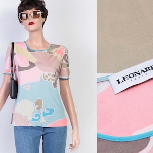 May include: A short-sleeved top with a colorful abstract pattern in pink, blue, beige, and brown. The top has a scoop neck and light blue trim. The tag reads "LEONARD PARIS". The top is paired with blue jeans.