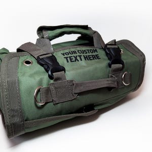May include: A green canvas roll-up bag with a black strap and buckle. The bag has a handle and a label that says "YOUR CUSTOM TEXT HERE".