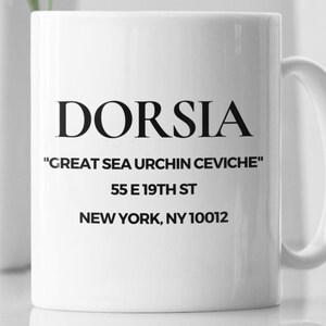 May include: White ceramic mug with the word "DORSIA" in bold black letters. Below, the text reads "GREAT SEA URCHIN CEVICHE" and an address. A simple, elegant design for a restaurant or cafe.