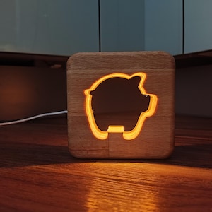 Könnte beinhalten: Ein Nachtlicht aus Holz in Form eines Sparschweins mit einem Ausschnittmuster. Das Licht ist eingeschaltet und strahlt ein warmes Licht aus.