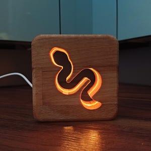 Könnte beinhalten: Ein Nachtlicht aus Holz in Form einer Schlange mit warmem orangefarbenem Leuchten. Das Licht ist eingesteckt und steht auf einer Holzoberfläche.