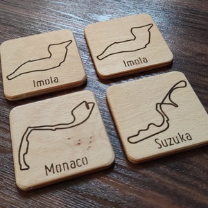 Könnte beinhalten: Vier quadratische Untersetzer aus Holz mit eingravierten Rennstrecken-Designs. Jeder Untersetzer zeigt eine andere Strecke: Imola, Monaco und Suzuka. Die Untersetzer sind aus hellem Holz gefertigt.