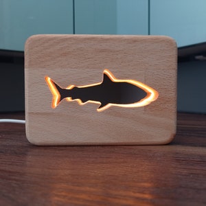 Puede incluir: Una luz nocturna de madera con forma de tiburón con un brillo cálido que emana del recorte. La luz está enchufada y sentada sobre una superficie de madera.
