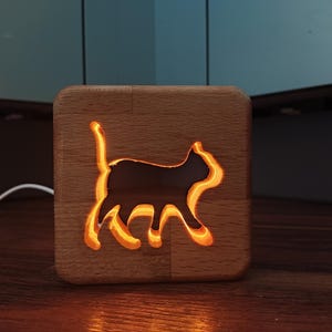 Puede incluir: Una luz de noche de madera con un recorte de silueta de gato. La forma del gato brilla con una luz naranja, dentro de un bloque de madera cuadrado. Un cable blanco se extiende desde la base. La luz está sobre una superficie de madera.