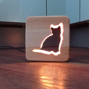 Puede incluir: Una luz nocturna de madera con forma de gato. El gato está recortado en la madera y la luz brilla a través del recorte. La luz nocturna está sobre una superficie de madera.