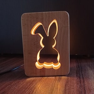 Puede incluir: Una luz nocturna de madera con forma de conejo con un brillo cálido que emana del recorte. La luz está sobre una superficie de madera.