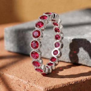 May include: A silver-toned eternity band featuring a continuous row of round, vibrant red gemstones. Each stone is individually set within a polished metal bezel, creating a uniform and circular design that encircles the entire circumference of the ring.
