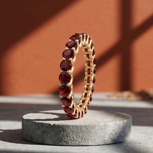 Könnte beinhalten: Ein goldener Ring mit einer durchgehenden Reihe runder, tiefroter Edelsteine. Der Ring wird auf einem kleinen, grauen Betonsockel vor einem warmen, orangebraunen Hintergrund präsentiert. Das Design des Rings ist klassisch und elegant.