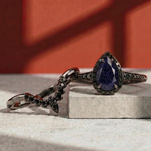 May include: Two rings: a black metal band with a curved design of black stones and a black metal ring with a pear-shaped blue stone. The blue stone has a speckled appearance and is surrounded by smaller black stones.