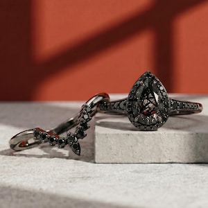 May include: Two black rings. One ring has a pear-shaped, dark grey gemstone surrounded by small black stones. The other ring is a curved band with multiple black stones. The rings are displayed on a white surface against a red background.