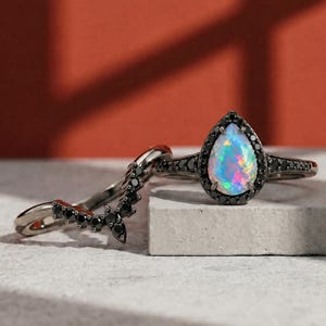 May include: A pear-shaped opal ring with a black halo setting and a matching curved band. The opal displays a spectrum of colours. The rings are set against a neutral background with a red backdrop.