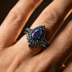 May include: A black ring set featuring a pear-shaped, dark blue gemstone with sparkling inclusions. The gemstone is surrounded by small, black accent stones. The ring is on a finger.