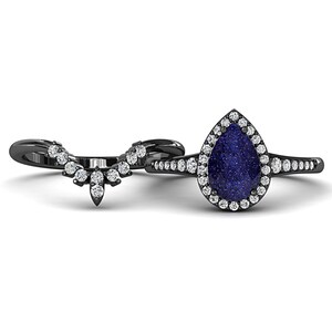 May include: A black metal ring set featuring a pear-shaped blue gemstone surrounded by small white stones. The matching band has a curved design with multiple white stones. The rings are set against a white background.