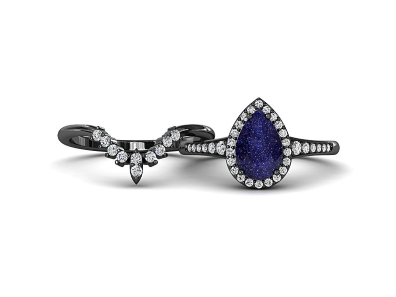 May include: A black metal ring set featuring a pear-shaped blue gemstone surrounded by small white stones. The matching band has a curved design with multiple white stones. The rings are set against a white background.