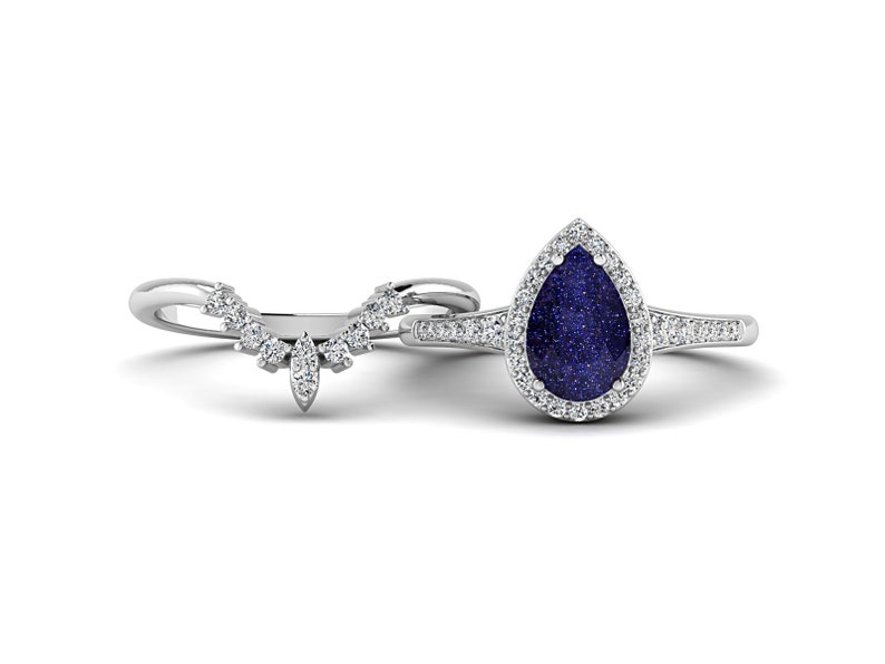 May include: A bridal ring set featuring a pear-shaped blue gemstone ring with a halo of small white stones and a matching curved band. The rings are set in a silver-colored metal, creating a classic and elegant look.