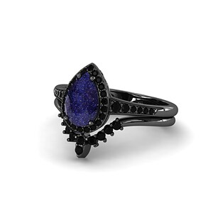 May include: A black ring set featuring a pear-shaped blue gemstone with a sparkling appearance, surrounded by smaller black stones. The band is black and includes additional black stones.