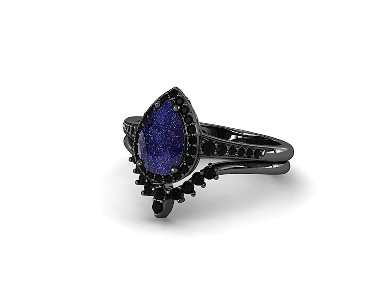 May include: A black ring set featuring a pear-shaped blue gemstone with a sparkling appearance, surrounded by smaller black stones. The band is black and includes additional black stones.