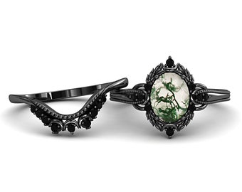 May include: A black engagement ring set. The main ring has an oval moss agate stone, and the set includes a curved band. The rings have black detailing and are displayed on a white backdrop.