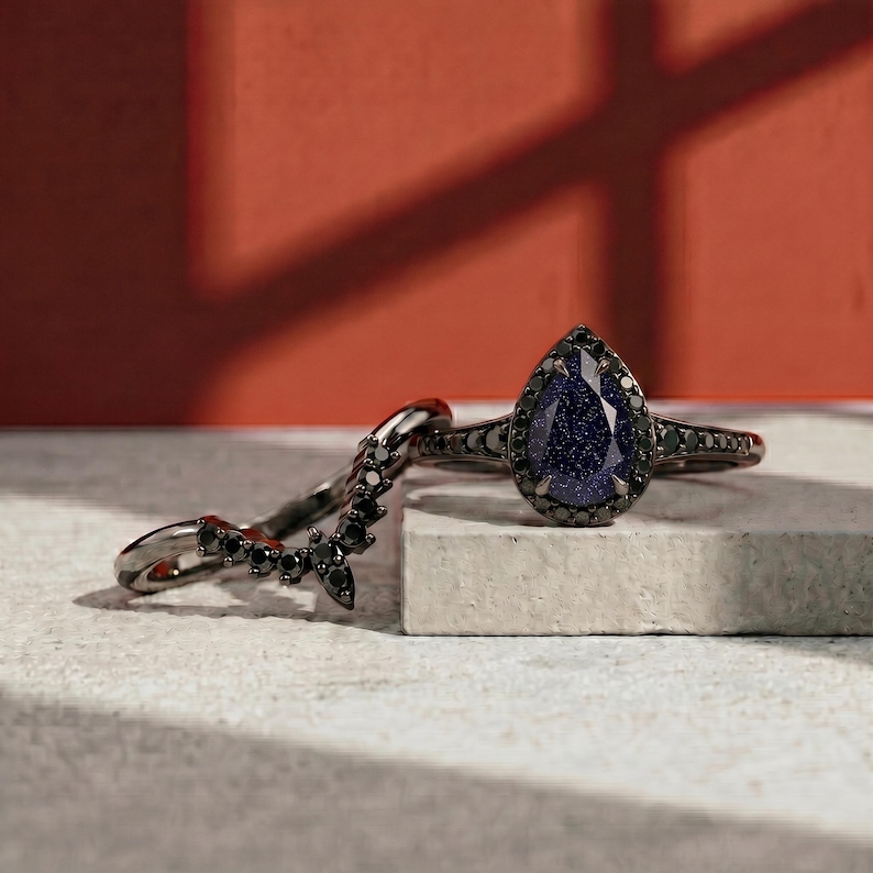 May include: Two rings: a black metal band with a curved design of black stones and a black metal ring with a pear-shaped blue stone. The blue stone has a speckled appearance and is surrounded by smaller black stones.