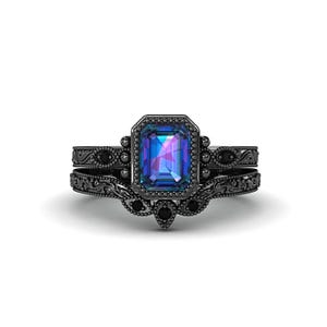 Könnte beinhalten: Ein schwarzes Verlobungsring-Set mit einem smaragdgeschliffenen, farbwechselnden Edelstein. Der Mittelstein ist von einem Halo aus kleineren Steinen umgeben. Das Band hat ein verziertes, filigranes Design mit zusätzlichen schwarzen Steinen. Das Set enthält ein passendes Band.
