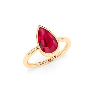 May include: A gold ring featuring a pear-shaped, faceted, bright red gemstone. The gemstone is bezel-set, and the ring band is a simple, polished gold. The ring is set against a white background.