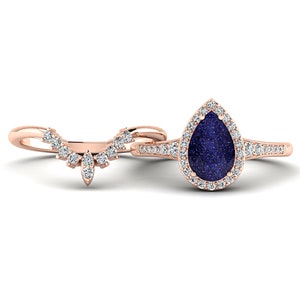 May include: A rose gold engagement ring set. The main ring features a pear-shaped, dark blue gemstone surrounded by a halo of small, clear stones. The band is also adorned with small stones. The accompanying band is curved and set with small, clear stones.