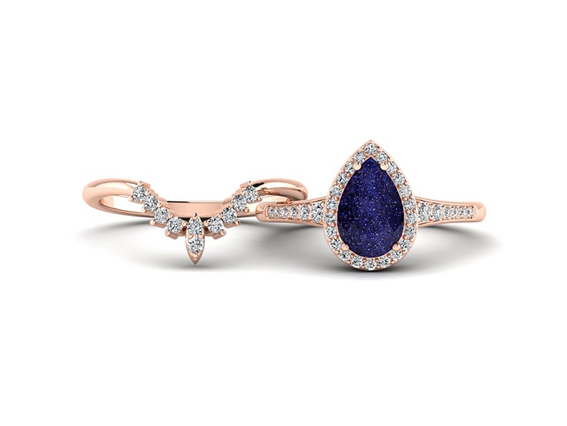 May include: A rose gold engagement ring set. The main ring features a pear-shaped, dark blue gemstone surrounded by a halo of small, clear stones. The band is also adorned with small stones. The accompanying band is curved and set with small, clear stones.