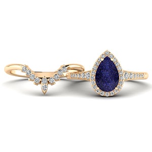 May include: A gold-colored engagement ring set. The primary ring features a pear-shaped, dark blue gemstone surrounded by a halo of small, clear stones. The accompanying band is a curved design with multiple small, clear stones.