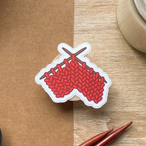 May include: A red knitted stitch sticker with two brown knitting needles. The sticker is on a brown surface with a roll of tape and a wooden surface.