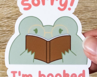 Op de afbeelding: Een witte sticker met een groene kikker die een bruin boek leest. De sticker zegt "sorry! Ik ben volgeboekt".