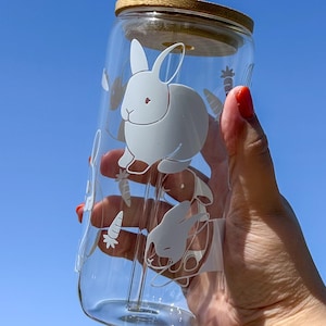 Peut inclure: Un gobelet en verre transparent avec un couvercle en bambou et une paille en verre transparent. Le gobelet est décoré d'illustrations blanches de lapins et de carottes. Le gobelet est tenu contre un ciel bleu vif.