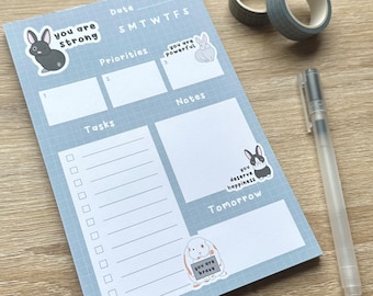 May include: A blue and white gridded notepad with a motivational quote and bunny illustrations. The notepad has sections for tasks, priorities, notes, and a calendar. The text on the notepad reads "you are strong", "you are powerful", "you deserve happiness", "you are brave", and "Tomorrow".