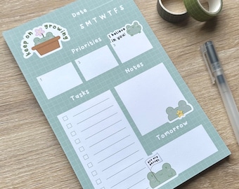 May include: A green and white daily planner with a grid pattern. The planner has sections for tasks, priorities, notes, and a calendar. It features frog illustrations with motivational messages like "Keep on growing" and "I believe in you!"