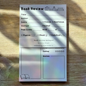 May include: A white book review notepad with a grid pattern and a black and white illustration of a book. The notepad has a title section for book title, author, genre, started, finished, page count, and a section for quotes. The notepad also has a section for rating the book with five stars.